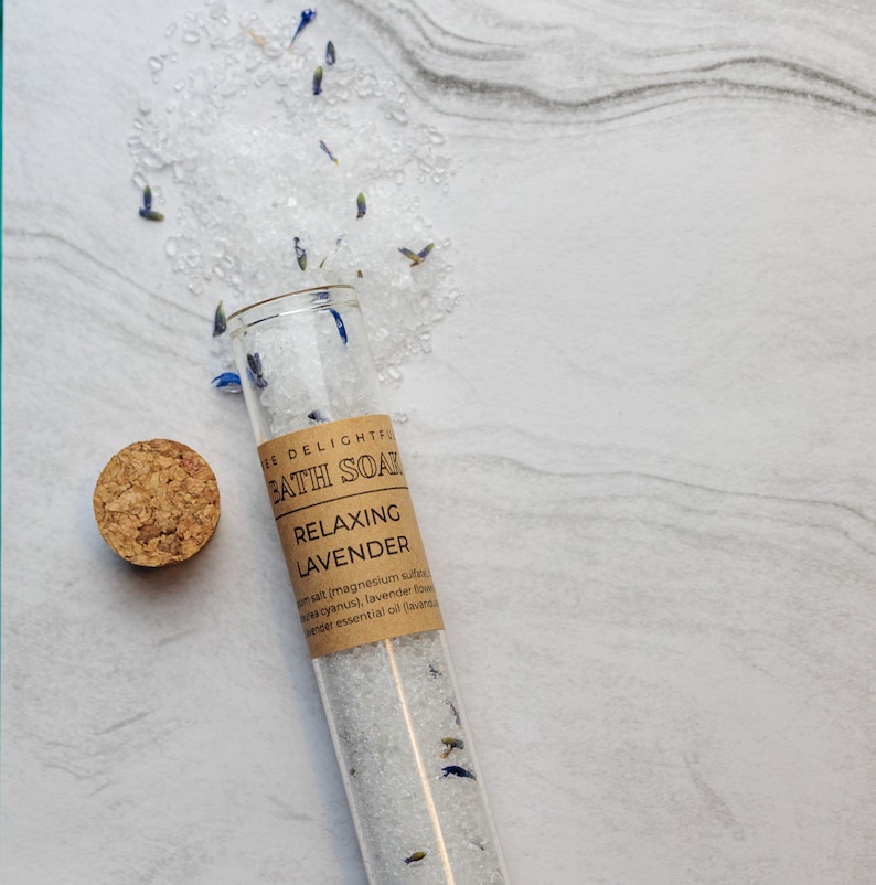 Puede incluir: Un tubo de vidrio lleno de sales de ba&ntilde;o blancas y flores de lavanda. La etiqueta dice "E DELIGHTFUL BATH SOAK RELAXING LAVENDER salt (magnesium sulfate), dried cyanus, lavender flowers, lavender essential oil (lavender"