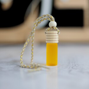 May include: A yellow glass bottle with a wooden cap and a string for hanging. The bottle is filled with a yellow liquid. The string is a light beige color with gold accents.