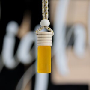 May include: A small, yellow glass bottle with a wooden cap and a string for hanging. The bottle is filled with a yellow liquid. The string is a beige color with gold accents. The background is blurred.