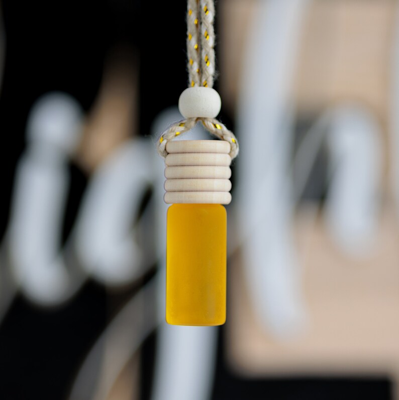 May include: A small, yellow glass bottle with a wooden cap and a string for hanging. The bottle is filled with a yellow liquid. The string is a beige color with gold accents. The background is blurred.