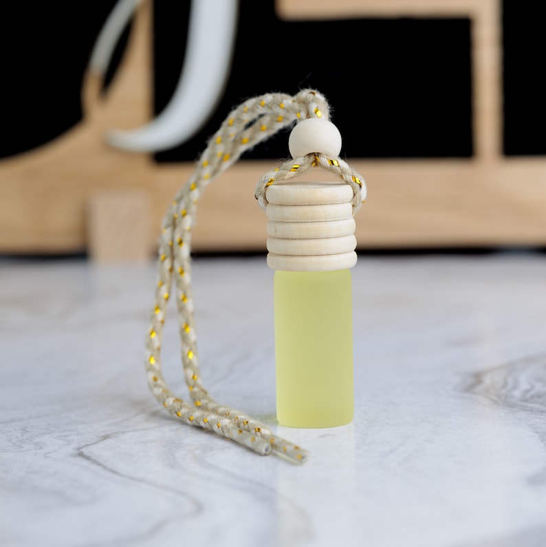 May include: A small, frosted glass bottle filled with a pale yellow liquid, topped with a wooden cap and a decorative rope. The rope is beige with gold accents. The bottle is designed to hang.