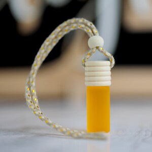 May include: A small, rectangular, yellow glass bottle with a wooden cap and a beige rope. The rope has gold flecks. The bottle is suspended by the rope. The background is blurred.