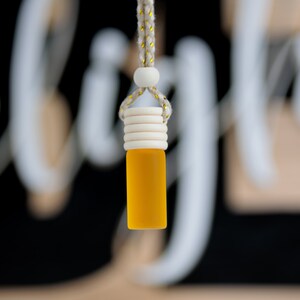 May include: A small, yellow glass bottle with a wooden cap and a woven rope for hanging. The bottle is filled with a yellow liquid. The background is blurred, with white and black elements.
