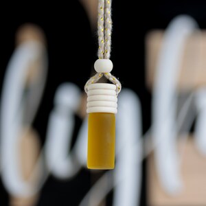 May include: A hanging car air freshener with a yellow liquid inside a small glass bottle. The bottle has a wooden cap and is suspended by a beige rope with gold accents. The background is black with blurred white and beige shapes.