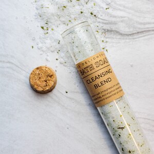 Puede incluir: Un tubo de vidrio lleno de sales de ba&ntilde;o blancas y hierbas verdes, etiquetado como "The Delightful Bath Soak Cleansing Blend" con una lista de ingredientes que incluyen sal de Epsom, sulfato de magnesio, menta, aceite esencial de eucalipto y mentol.