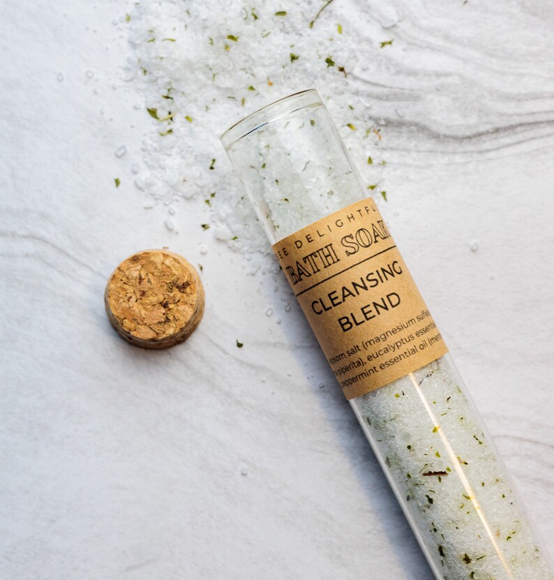 Puede incluir: Un tubo de vidrio lleno de sales de ba&ntilde;o blancas y hierbas verdes, etiquetado como "The Delightful Bath Soak Cleansing Blend" con una lista de ingredientes que incluyen sal de Epsom, sulfato de magnesio, menta, aceite esencial de eucalipto y mentol.