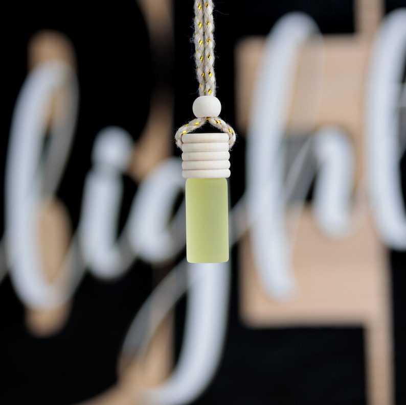 May include: A small, rectangular, light green glass bottle with a wooden cap and a beige rope for hanging. The bottle is suspended against a blurred black and white background. The rope has gold accents.