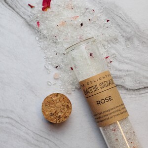 Puede incluir: Un tubo de vidrio lleno de sales de ba&ntilde;o blancas y p&eacute;talos de rosa. El tubo tiene una etiqueta de papel marr&oacute;n que dice "Jee Delightful Bath Soak Rose Epsom salt (magnesium sulfate), rose petals (rosa centifolia), vitamin E oil, essential oil (rosa damascena)"