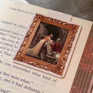 Peut inclure: Un marque-page rectangulaire présentant une reproduction imprimée d'une peinture classique dans un cadre orné aux tons métalliques. L'œuvre représente deux personnages en tenue historique dans un décor architectural en pierre, posé dans un livre ouvert.