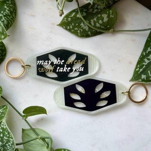 May include: Two clear plastic keychains with gold rings. One keychain has a black background with the text "may the dread wolf take you" in gold lettering. The other keychain has a black background with a leaf design.