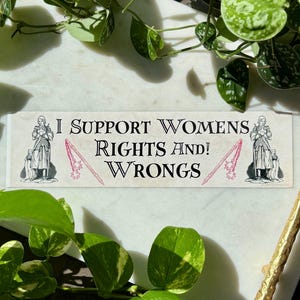 May include: A rectangular sticker with the text "I SUPPORT WOMENS RIGHTS AND! WRONGS" in black, with two illustrations of armored figures and pink maces. The sticker is on a white surface, surrounded by green plants.
