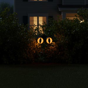 May include: A pair of glowing, amber-colored cat eyes with vertical black pupils, mounted in a black frame, positioned within a dark green hedge at night, emitting a warm light that illuminates the surrounding leaves and foliage.
