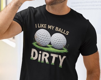 May include: Black t-shirt with the text "I LIKE MY BALLS DIRTY" in white and green. The graphic features two golf balls on a patch of green grass. The shirt is worn by a person.