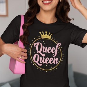 Puede incluir: Camiseta negra con el texto "Queef Queen" en rosa, rodeada por un círculo dorado y estrellas, coronada con una corona dorada. La camiseta se combina con pantalones cortos blancos y un bolso rosa.