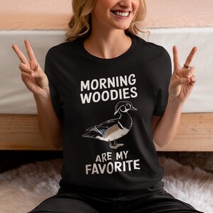 May include: A black t-shirt with white text that reads "Morning Woodies are my favorite". A cartoon image of a wood duck is printed on the shirt.