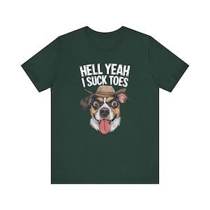 Puede incluir: Una camiseta de manga corta de color verde oscuro con el gráfico de un perro que lleva un sombrero de vaquero marrón y tiene la lengua fuera. Sobre el perro, el texto blanco HELL YEAH I SUCK TOES aparece en una fuente negrita y desgastada.