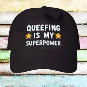May include: Black baseball cap with white embroidered text that reads "Queefing is my superpower" with two yellow stars on either side of the word "my".
