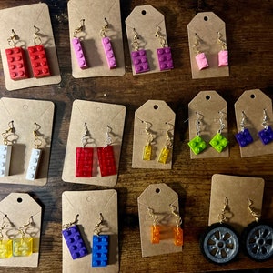 May include: A collection of 10 pairs of earrings featuring miniature Lego bricks in various colors, including red, blue, green, yellow, white, and purple. The earrings are displayed on small cards with white backgrounds.