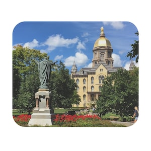 May include: A scenic view of a university campus featuring a statue in the foreground and a large building with a gold dome in the background. The building is surrounded by trees and red flowers under a blue sky.