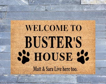 May include: A brown doormat with the text "Welcome to Buster's House" and two black paw prints. The text "Matt & Sara Live here too." is printed below the main text.
