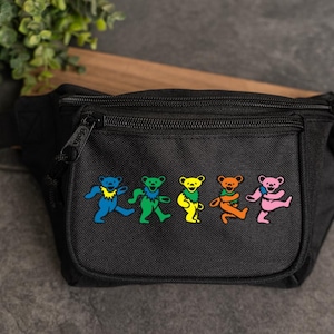 May include: Black fanny pack with a zipper closure and a colorful graphic of dancing bears. The bears are in blue, green, yellow, orange, and pink. The fanny pack is made of a durable material and has an adjustable strap.