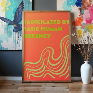 May include: Framed art print with the text "RADICILAZED BY BASIC HUMAN DECENCY" in green on an orange background. The print features green wavy lines and is displayed on a wooden floor. Other abstract art pieces are visible in the background.