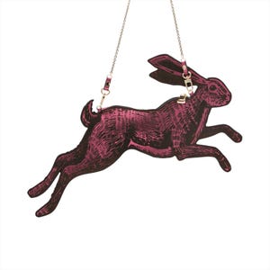 May include: A magenta and black crossbody bag shaped like a running hare. The bag has a detailed illustration of the hare and is suspended by a silver chain. The bag has a leather strap and a metal clasp.