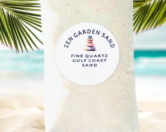 Può includere: Una bustina bianca con un'etichetta circolare bianca che dice "Zen Garden Sand" con un cumulo di pietre colorate al centro. L'etichetta dice anche "Fine Quartz Gulf Coast Sand". La bustina è appoggiata su una spiaggia di sabbia bianca.