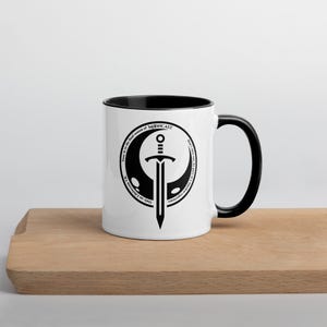May include: White ceramic mug with a black rim and handle. The mug features a black and white graphic of a sword and crescent moon with the text "Time is the first season of MOONCAST. Don't know what to do? Choose your adventure. Cook or Fight?"
