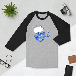 May include: A grey and black baseball tee shirt with a blue and white cartoon shark illustration wearing a pirate hat and holding a sword. The text "Ahoy" is written next to the shark.