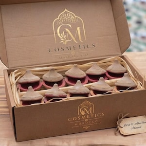 Peut inclure: Une boîte en carton brun contenant dix petits pots cosmétiques en terre cuite en forme de cône avec couvercles, nichés dans de la paille. Le couvercle et l'avant portent un logo doré avec le texte CM COSMETICS MOROCCO. Une étiquette indique Lot de 10-Aker Fassi Artisanal.