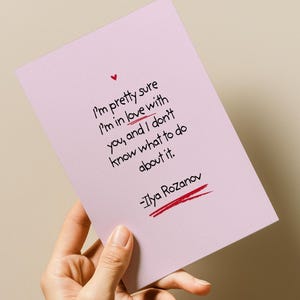 May include: A light pink greeting card with a handwritten quote in black ink: "I'm pretty sure I'm in love with you, and I don't know what to do about it." A small red heart and Ilya Rozanov's signature are also present.
