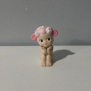 Pu&ograve; includere: Piccola statuina di un personaggio simile a una pecora con viso, orecchie e capelli rosa. La statuina ha un'espressione dolce ed &egrave; in posa con le mani giunte. La pecora ha un fiore tra i capelli ed &egrave; seduta.