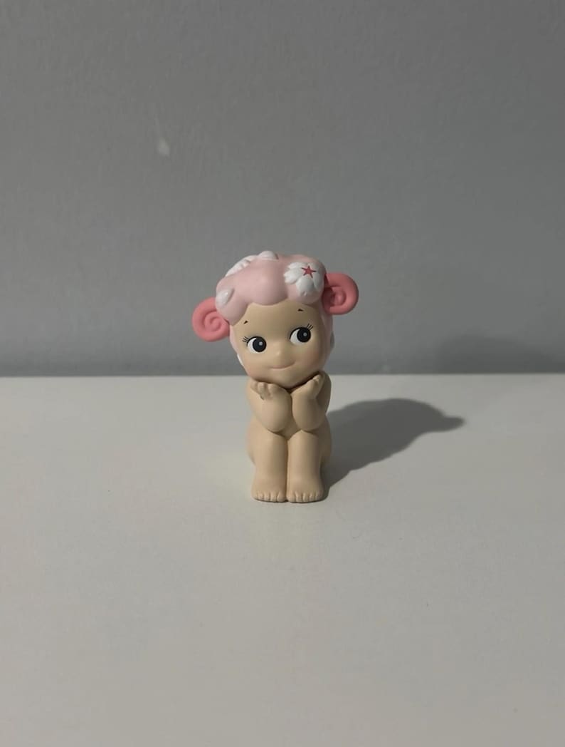 Pu&ograve; includere: Piccola statuina di un personaggio simile a una pecora con viso, orecchie e capelli rosa. La statuina ha un'espressione dolce ed &egrave; in posa con le mani giunte. La pecora ha un fiore tra i capelli ed &egrave; seduta.