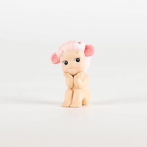 Pu&ograve; includere: Piccola statuina di un personaggio simile a una pecora, con capelli, orecchie e un fiore rosa. La figura &egrave; seduta con le mani vicino al viso e ha grandi occhi espressivi. Il design &egrave; carino e stravagante.