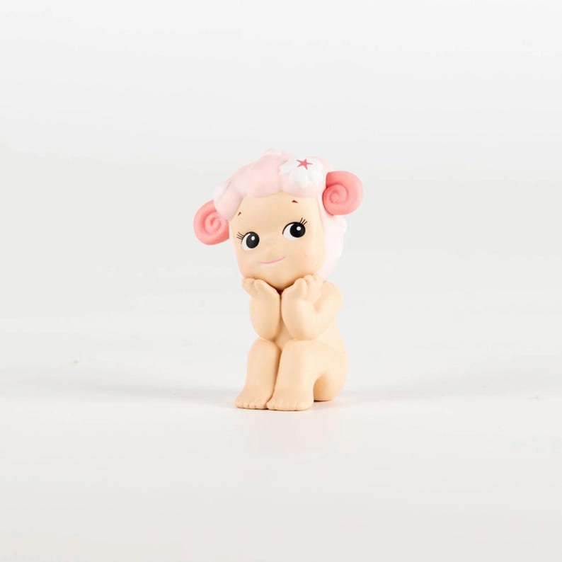 Pu&ograve; includere: Piccola statuina di un personaggio simile a una pecora, con capelli, orecchie e un fiore rosa. La figura &egrave; seduta con le mani vicino al viso e ha grandi occhi espressivi. Il design &egrave; carino e stravagante.