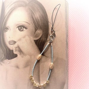 May include: A beaded phone charm with a black cord, silver-tone clasp, and a decorative strand. The strand features light blue beads, cream-coloured oval beads, and gold-coloured flower-shaped beads. The charm is displayed against a book page.