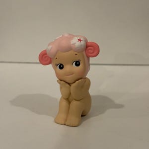 Pu&ograve; includere: Piccola statuina di un personaggio simile a una pecora con testa e orecchie rosa. La pecora ha un viso sorridente, occhi neri ed &egrave; in posa con le mani vicino al viso. La testa della pecora &egrave; adornata da un fiore.