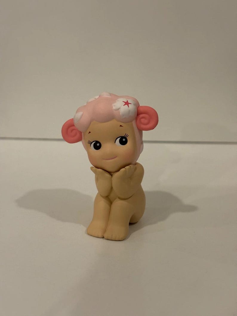 Pu&ograve; includere: Piccola statuina di un personaggio simile a una pecora con testa e orecchie rosa. La pecora ha un viso sorridente, occhi neri ed &egrave; in posa con le mani vicino al viso. La testa della pecora &egrave; adornata da un fiore.