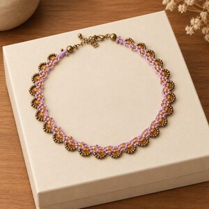 May include: A handcrafted necklace featuring light purple knotted cord woven with small amber-colored beads and metallic bronze-toned accents. The circular design includes a bronze-toned lobster claw clasp and an adjustable chain extension.