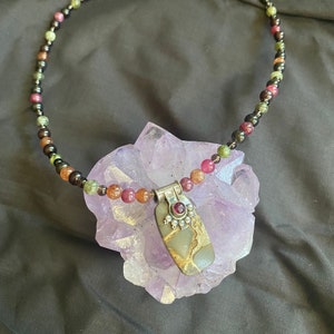 Peut inclure: Un collier composé de perles noires, vertes et roses avec un pendentif en argent orné d'une pierre brune et dorée et d'une pierre rouge. Le collier est présenté sur un cristal violet.