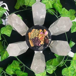 Puede incluir: Una colección de seis puntas de cristal de cuarzo claro dispuestas en círculo alrededor de un pequeño cuenco lleno de hierbas y flores secas. El cuenco es negro con un borde blanco.