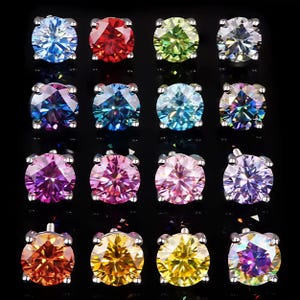 May include: A collection of twelve stud earrings with round-cut gemstones in various colors, including blue, red, green, and purple. Each earring is set in a silver-colored metal setting, displayed against a black background.