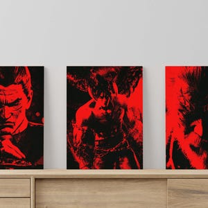 Può includere: Tre stampe artistiche incorniciate con ritratti stilizzati in rosso e nero. I soggetti includono un uomo, una figura con le corna e una creatura con occhi luminosi. L'opera d'arte è esposta su un mobile in legno.