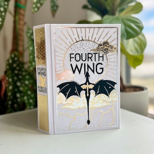 Könnte beinhalten: Ein Hardcover-Buch mit dem Titel "Fourth Wing" von Rebecca Yarros. Das Cover zeigt eine schwarze Drachensilhouette auf weißem Hintergrund mit goldenen Akzenten und dem Buchtitel in schwarzen Buchstaben.