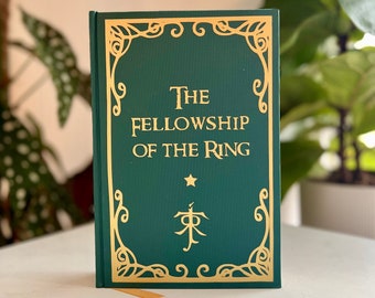 May include: A hardback book titled "The Fellowship of the Ring" with gold lettering and a decorative border on a dark green cover. The book features a star and a symbol. The book is displayed on a white surface.