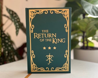 May include: A dark green hardcover book with the title "The Return of the King" in gold lettering. The cover has gold filigree and three gold stars. A gold ribbon bookmark is visible.