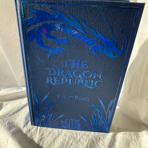 May include: A blue hardback book with a silver dragon design on the front cover. The title "The Dragon Republic" and the author "R.F. Kuang" are printed in silver lettering.