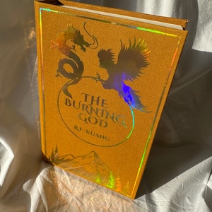 May include: A gold hardback book with a holographic cover featuring a dragon and a phoenix. The title of the book is "The Burning God" by R.F. Kuang.