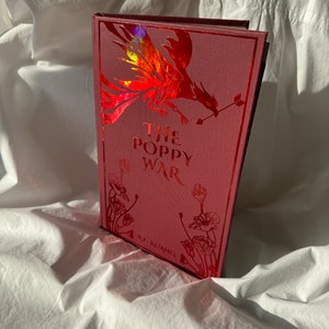 May include: A red hardback book with a holographic phoenix design on the cover. The title "The Poppy War" is printed in white lettering on the cover, with the author's name "R.F. Kuang" printed below the title.